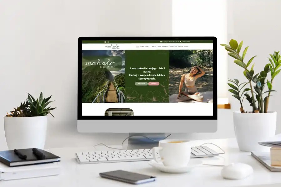 Ekran iMac z zieloną stroną internetową Mahalo Body Studio, na białym biurku z roślinami, kawą i akcesoriami. Minimalistyczny design, naturalne światło dzienne.