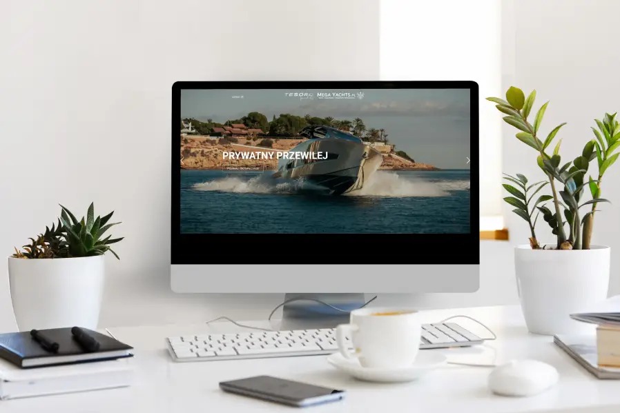 Ekran komputera stacjonarnego wyświetla stronę internetową z luksusowym jachtem na tle błękitnego morza; minimalistyczne biurko z roślinami, kawa i akcesoria.