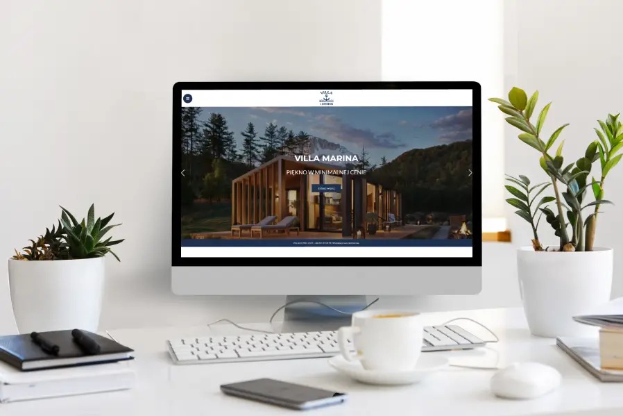 Monitor wyświetla stronę internetową Villa Marina z minimalistycznym domem na tle lasu. Na biurku roślina doniczkowa i filiżanka kawy.