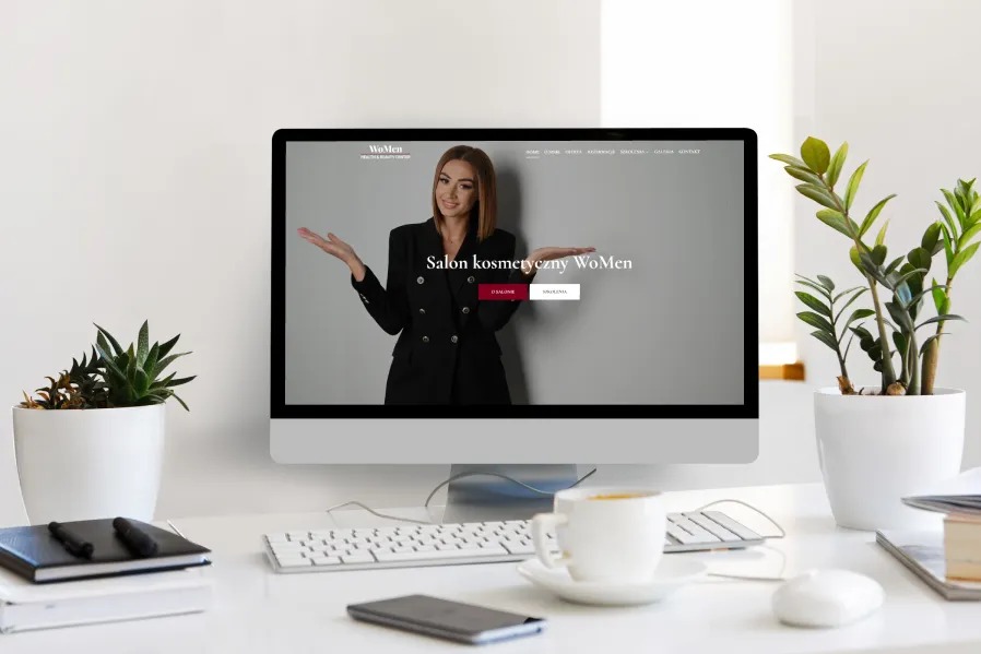 Ekran komputera stacjonarnego prezentuje stronę internetową salonu kosmetycznego WoMen, widoczny na blacie biurka z akcesoriami i roślinami doniczkowymi.