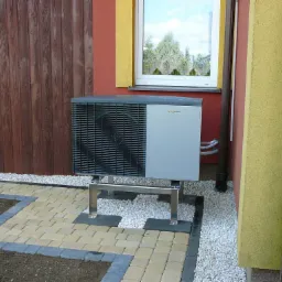 Zewnętrzna jednostka pompy ciepła Viessmann na metalowym stelażu, obok drewnianego ogrodzenia i kostki brukowej.