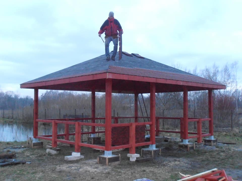 Czerwona altana z drewnianą konstrukcją i robotnikiem na dachu, w tle widoczne drzewa i małe jeziorko.