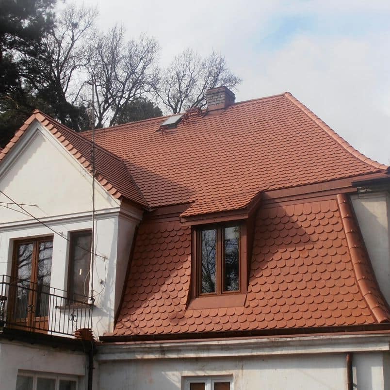 Wykończony dach z dachówki łuskowej w odcieniach czerwieni na dwupiętrowym domu z białymi ścianami, widoczny komin i okno dachowe.