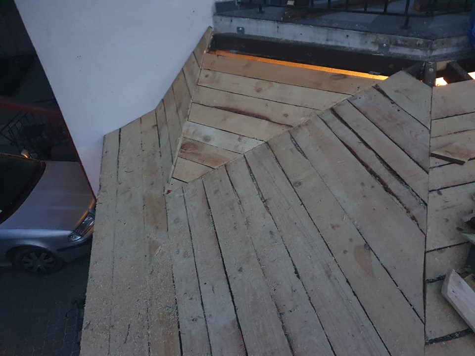 Surowe deski tworzące zarys konstrukcji, prawdopodobnie podłoże pod taras, widoczne elementy mocujące i fragment samochodu w tle.