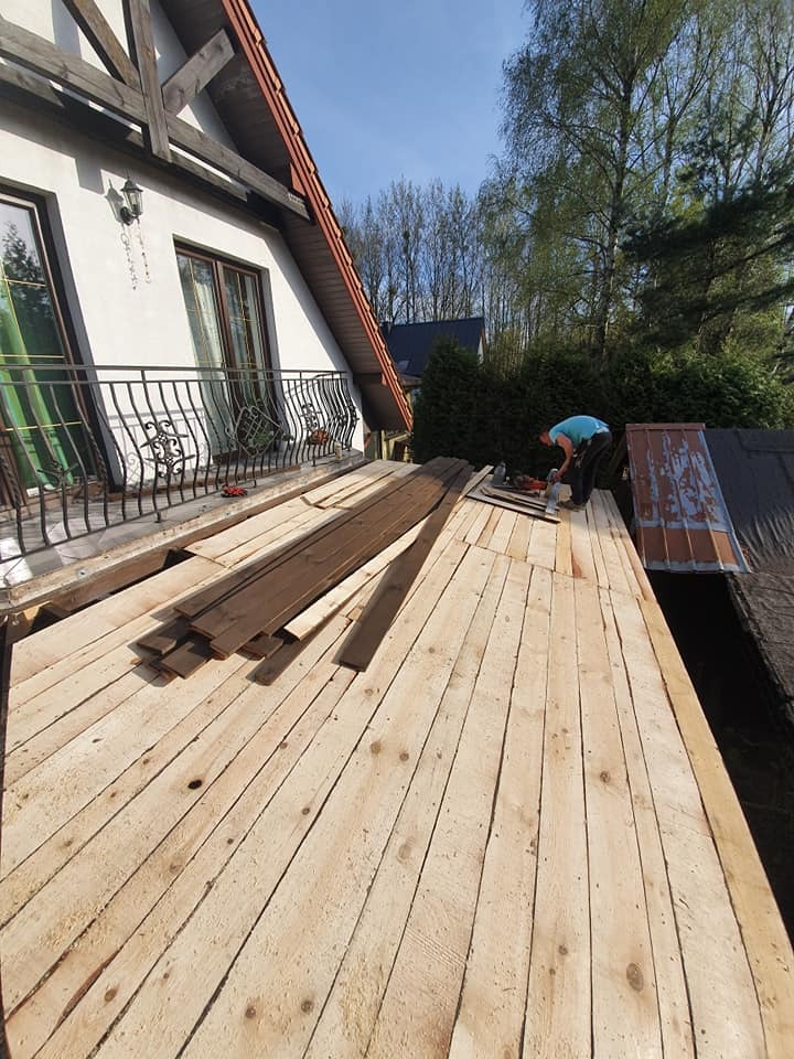 Prace dekarskie w toku: robotnik w niebieskiej koszulce montuje deski na dachu domu jednorodzinnego z balkonem, widoczne narzędzia i stare elementy dachu.