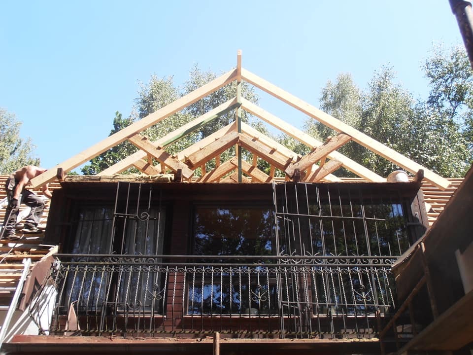 Prace dekarskie w toku: montaż drewnianej konstrukcji dachu nad balkonem z ozdobną, metalową balustradą i oknami z kratami. Widoczny dekarz na drabinie.