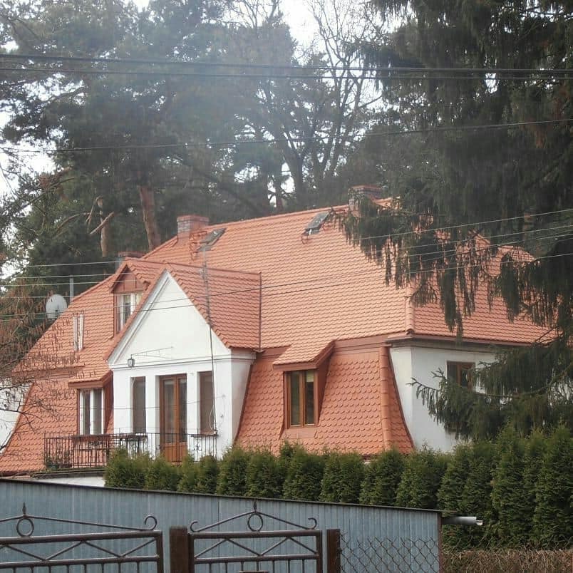 Dom jednorodzinny z nowym dachem z czerwonej dachówki, widoczny komin i obróbki blacharskie, otoczony zielenią, sfotografowany zza metalowego ogrodzenia.