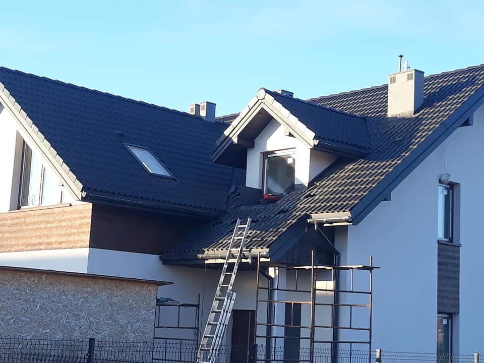 Dom jednorodzinny w trakcie prac dekarskich, widoczne rusztowania, drabina oparta o dach pokryty ciemną dachówką, okno dachowe i fragmenty obróbek blacharskich.