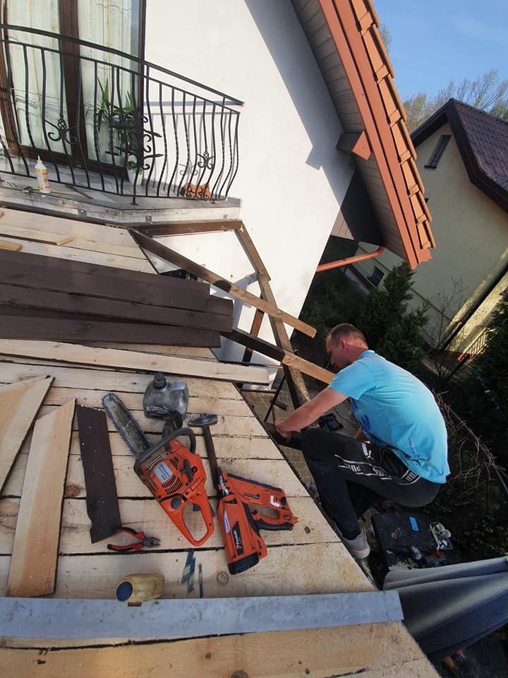 Pracownik w trakcie montażu pokrycia dachowego, widoczne narzędzia: pilarka spalinowa, gwoździarka pneumatyczna, obok leżą deski i blacha. W tle balkon z ozdobną balustradą i fragmenty sąsiednich...