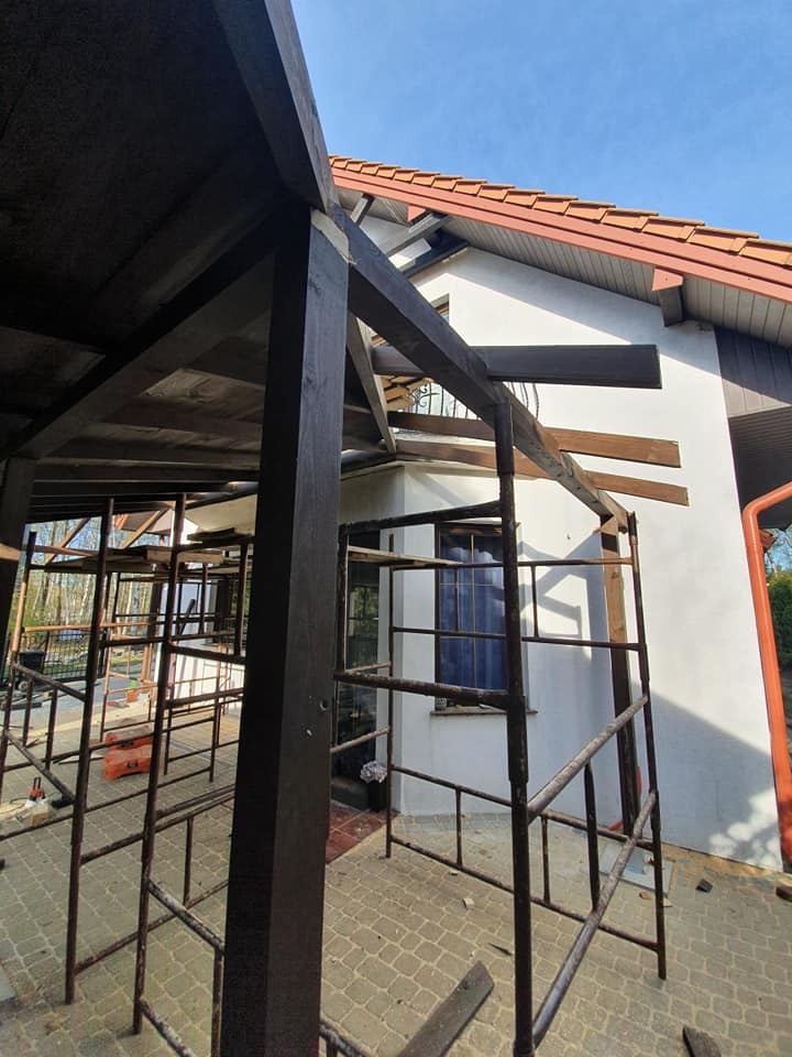 Konstrukcja z ciemnego drewna, prawdopodobnie zadaszenia, w trakcie montażu przy białym domu z czerwoną dachówką, widoczne rusztowania i fragmenty narzędzi.