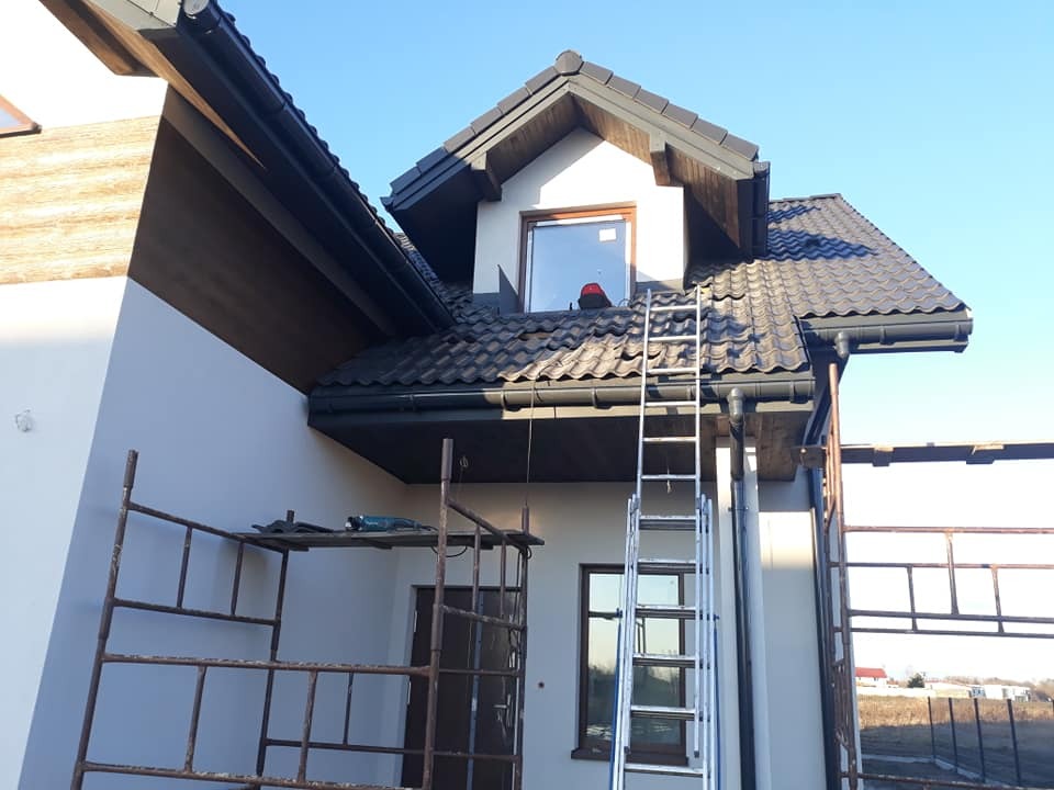 Dom jednorodzinny w trakcie montażu rynien i obróbek blacharskich, widoczne rusztowania i drabina oparta o dach z blachodachówki.