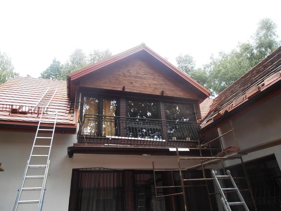 Dom w trakcie remontu dachu z widoczną drewnianą nadbudówką z balkonem, drabiny opierają się o budynek, częściowo zdjęte dachówki.