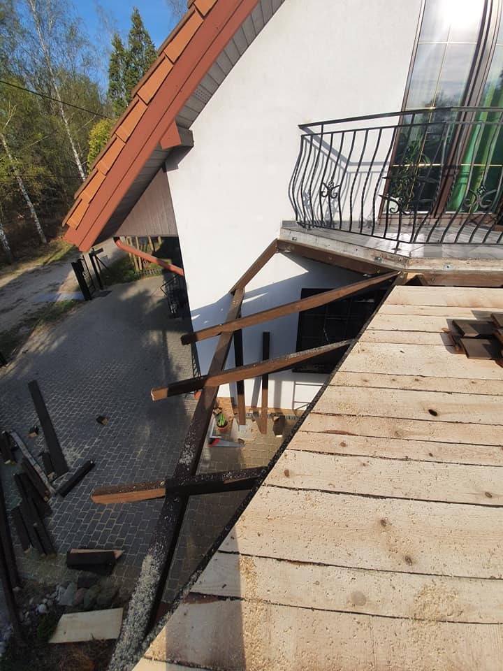 Budowa drewnianej konstrukcji dachu z widoczną więźbą, fragmentem istniejącego dachu z czerwonej dachówki i balkonem z ozdobną, metalową balustradą w tle.