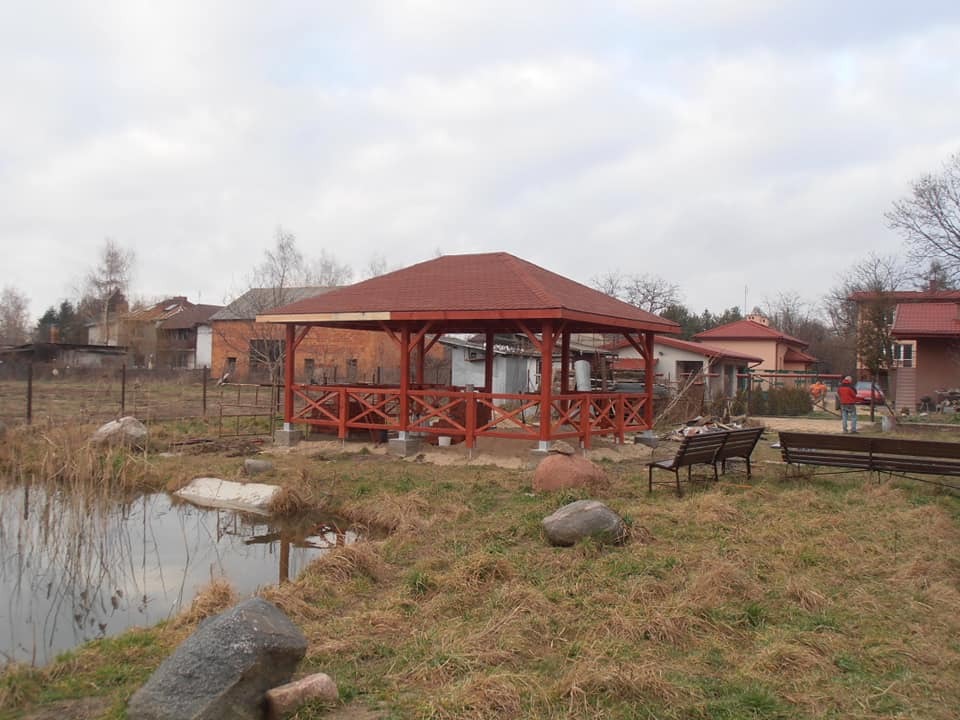 Nowa, pomalowana na czerwono altana z drewnianą balustradą, zadaszona czerwoną dachówką, stoi na betonowych fundamentach obok stawu w wiejskim otoczeniu.