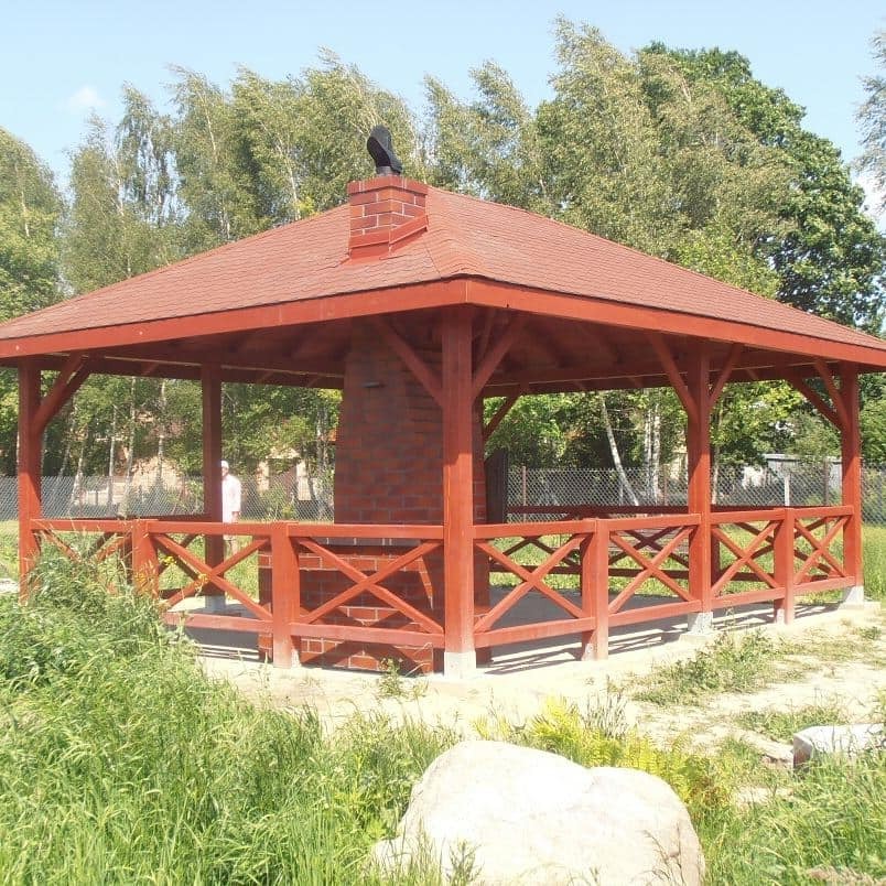 Czerwona, drewniana altana z ceglanym kominem i dachem z dachówki bitumicznej, otoczona zielenią i dużym kamieniem.