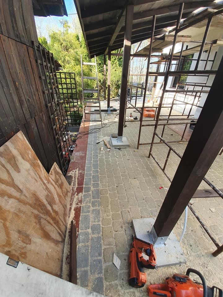 Budowa zadaszenia tarasu: widoczne drewniane słupy na betonowych podstawach, metalowa konstrukcja dachu w trakcie montażu, narzędzia budowlane na kostce brukowej, pionowa drabina oparta o ścianę...