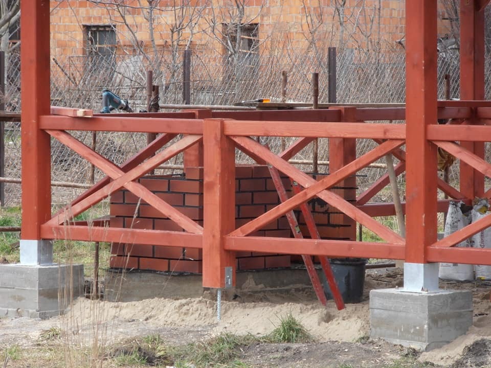 Konstrukcja drewnianej pergoli w trakcie budowy, widoczne ceglane elementy, betonowe podstawy, narzędzia i materiały budowlane w tle.