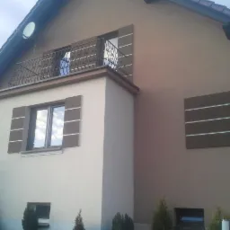 Dwukondygnacyjny dom z balkonem i dekoracyjnymi panelami elewacyjnymi w odcieniach beżu i brązu. Widoczne okna z dekoracyjnymi elementami i fragment schodów.