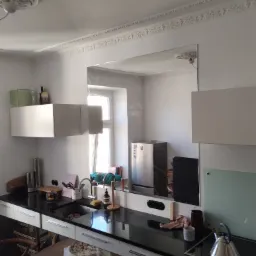 Duże lustro w kuchni, zamontowane na ścianie między szafkami, odbija fragment okna i lodówki; na pierwszym planie blat z czarnym kamieniem i białe szafki, na stole leżą narzędzia.