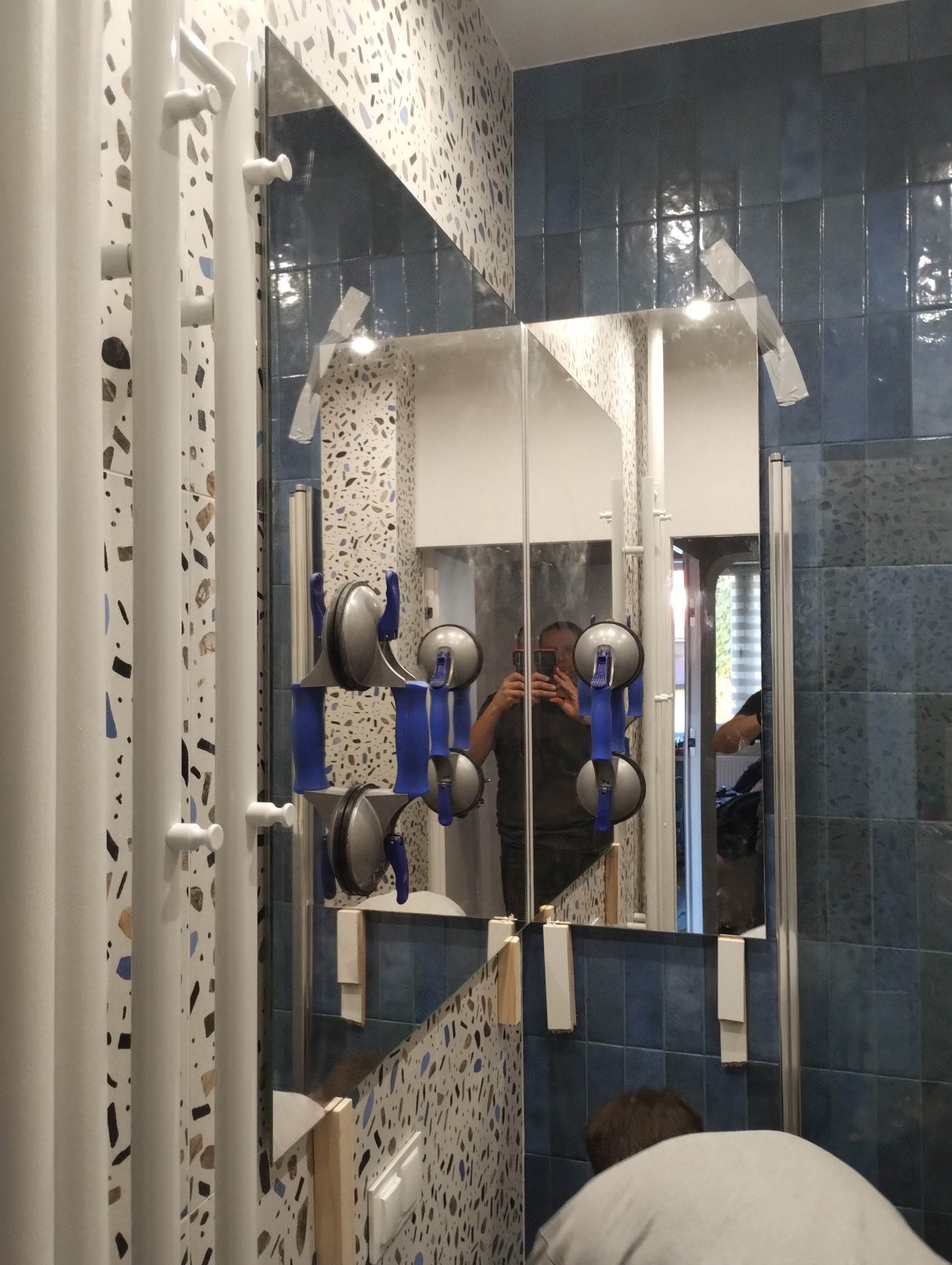 Montaż lustra w łazience z użyciem przyssawek, widoczne niebieskie uchwyty, fragment osoby robiącej zdjęcie w odbiciu, ściana wykończona płytkami w dwóch wzorach.