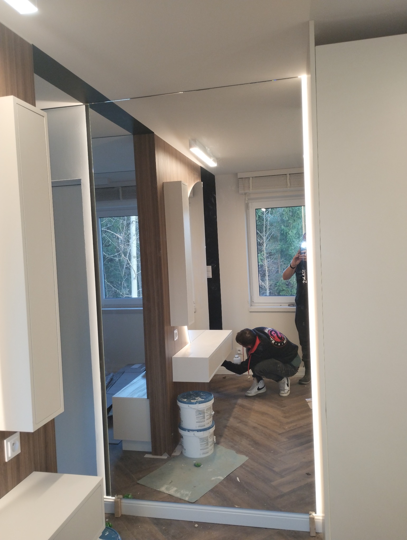 Instalacja lustra z podświetleniem LED w łazience, widoczne wiaderka z klejem i pracownik w trakcie montażu.