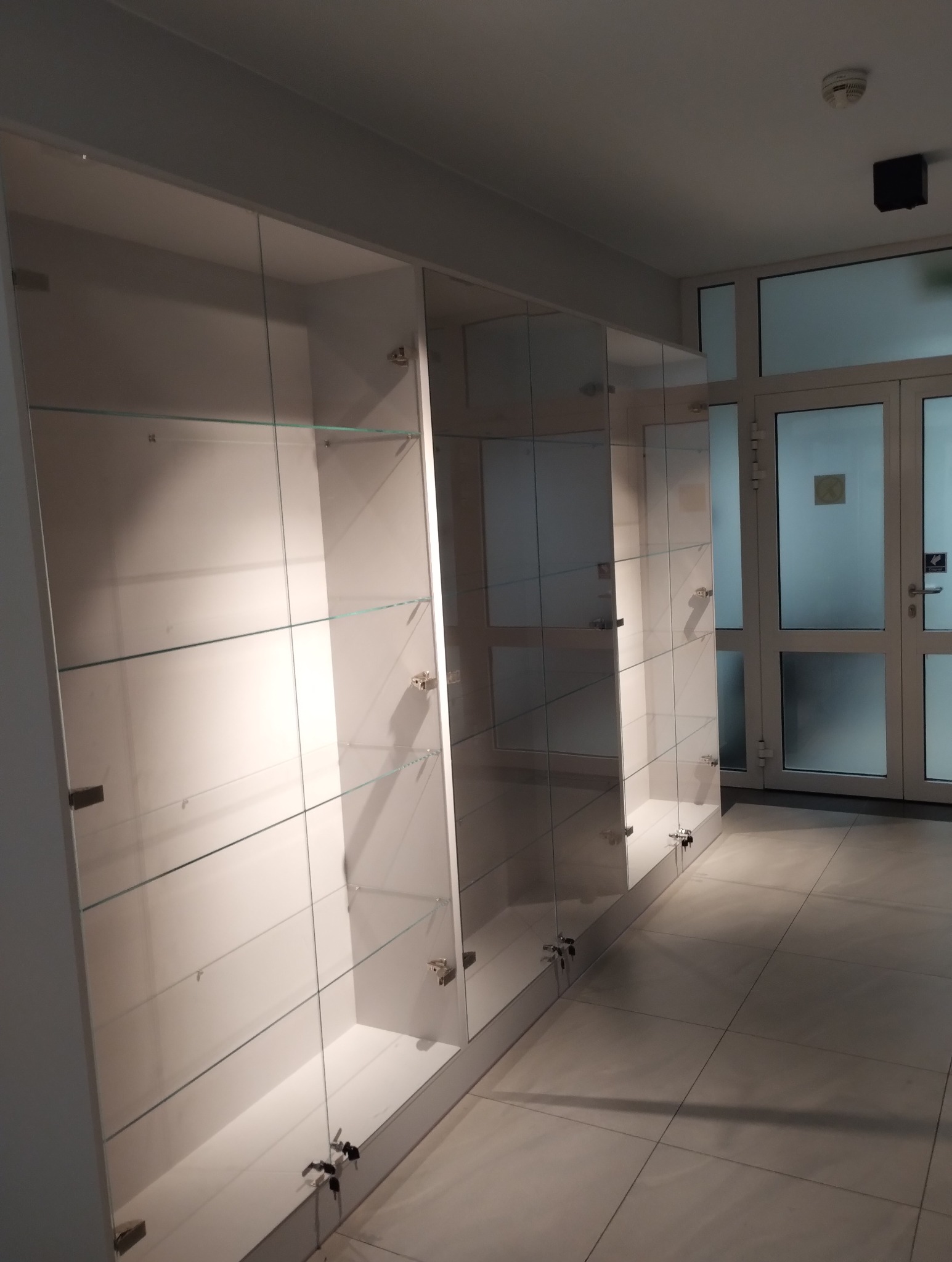 Długi rząd białych, przeszklonych witryn sklepowych z zamkami, ustawionych wzdłuż korytarza z białymi drzwiami i szarą podłogą z płytek.