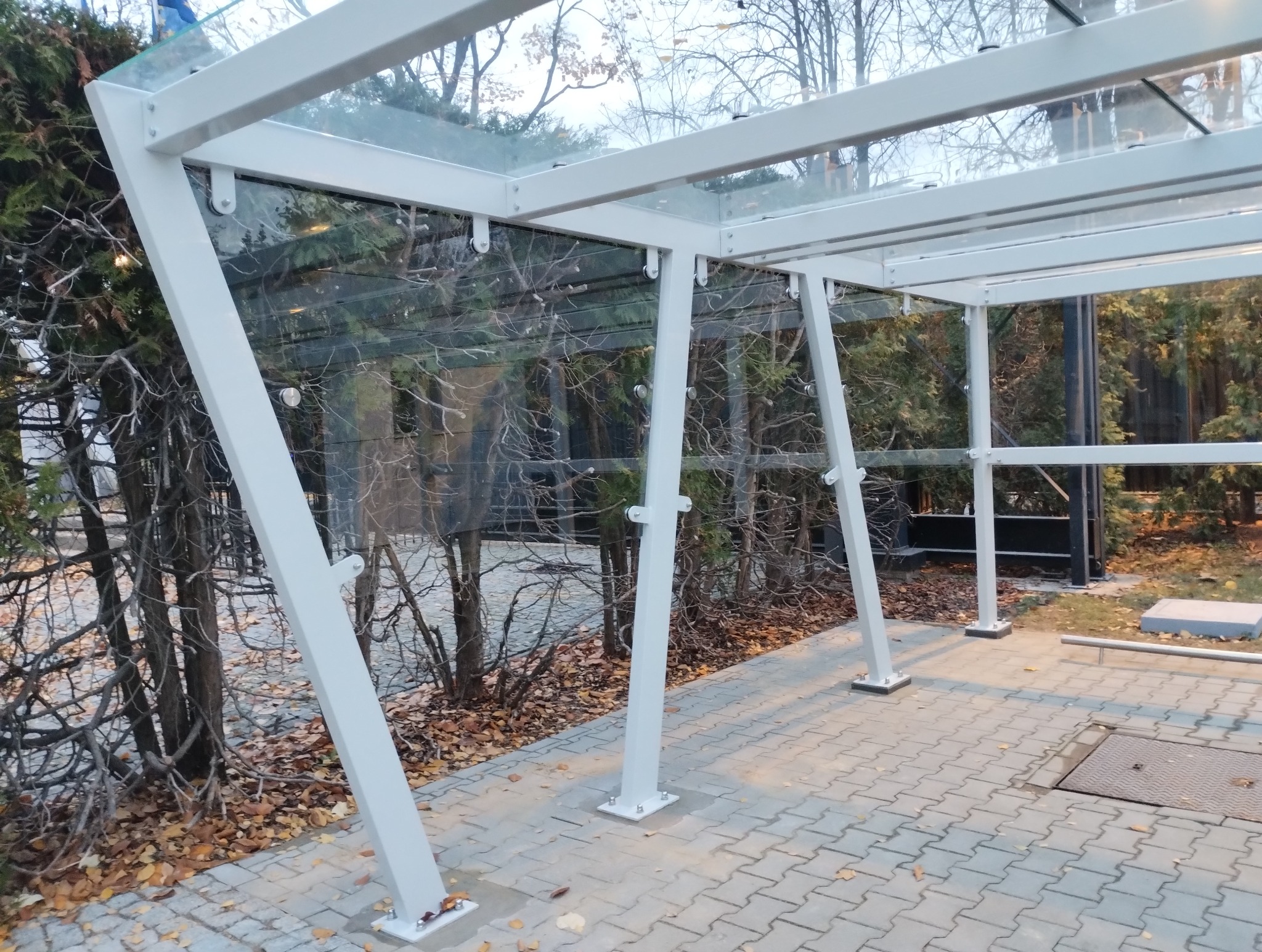 Zadaszenie szklane z białymi, ukośnymi podporami na zewnątrz, widoczne drzewa w tle.