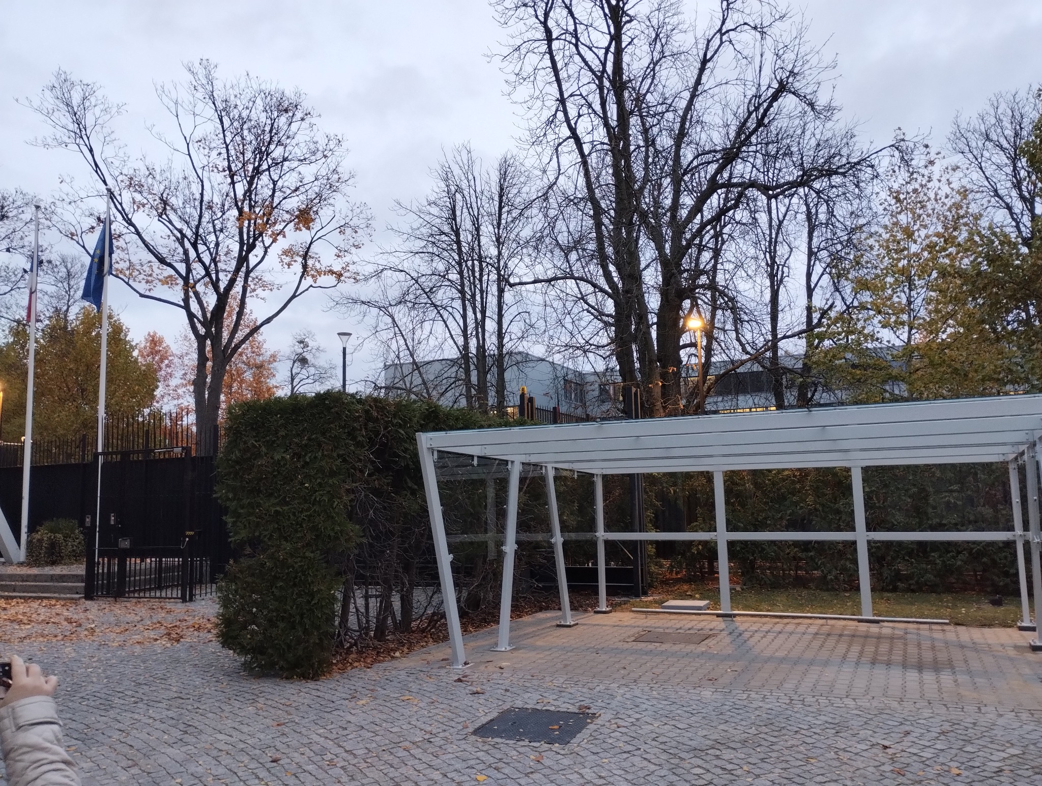 Zewnętrzny widok na przeszkloną wiatę przystankową z metalową konstrukcją, umieszczoną na brukowanym placu, z widocznymi drzewami i flagami w tle, fotografowaną z niskiego kąta.