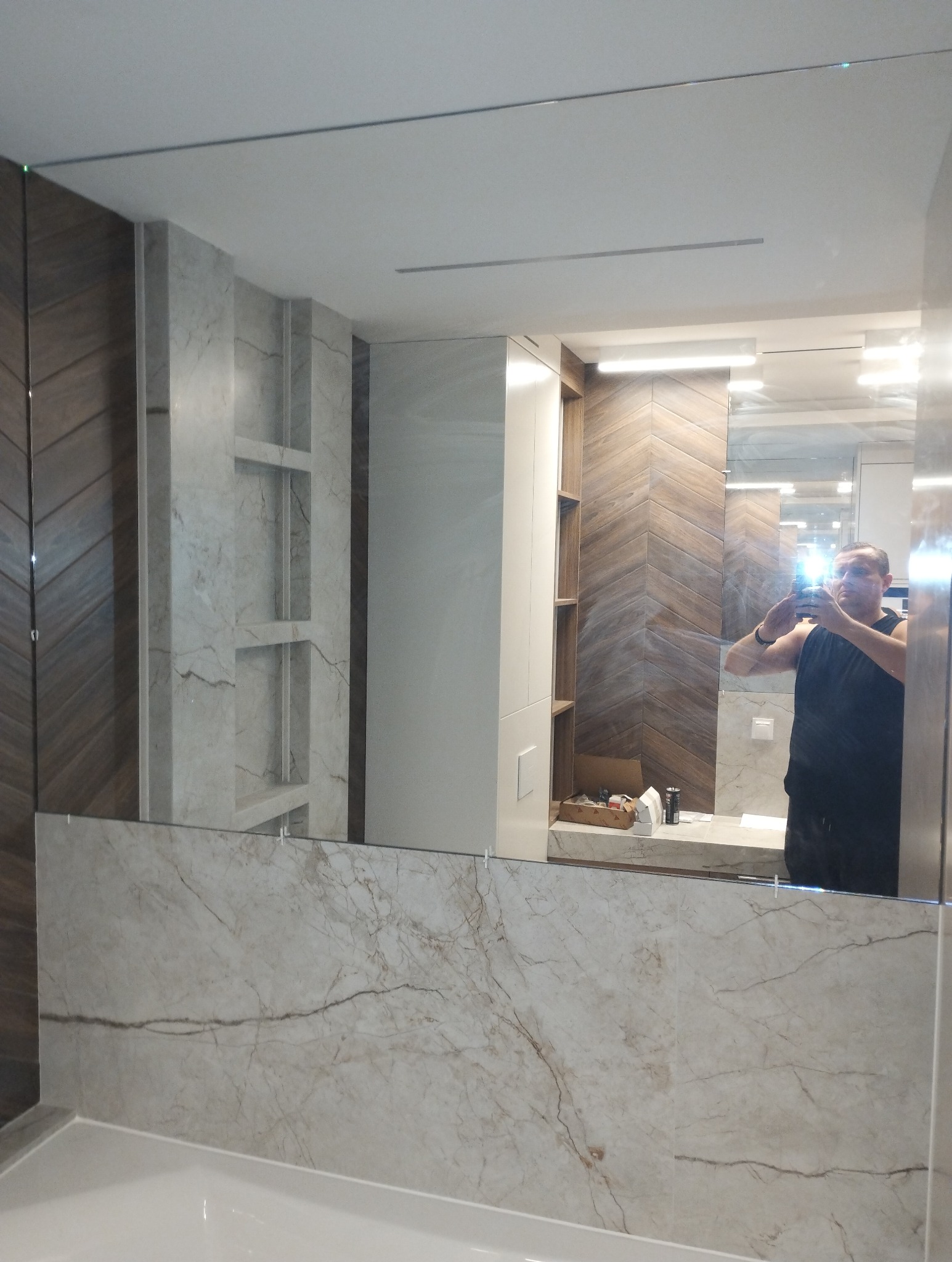 Nowoczesna łazienka z dużym lustrem na ścianie, odbicie mężczyzny robiącego zdjęcie telefonem, widoczne płytki imitujące marmur i drewno ułożone w jodełkę.
