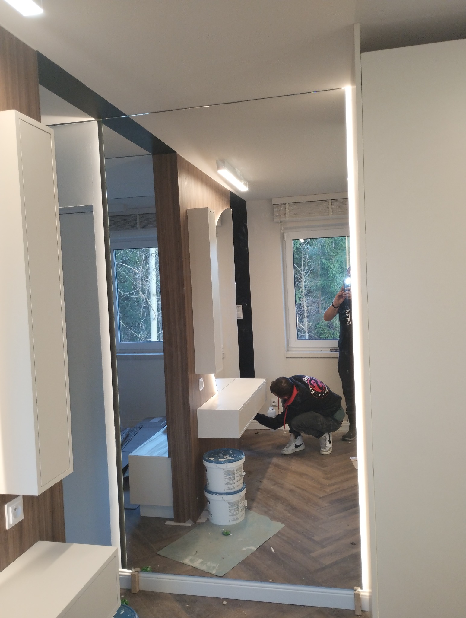 Montaż lustra z oświetleniem LED w łazience, widoczne dwie puszki z klejem i osoba pracująca nad instalacją.