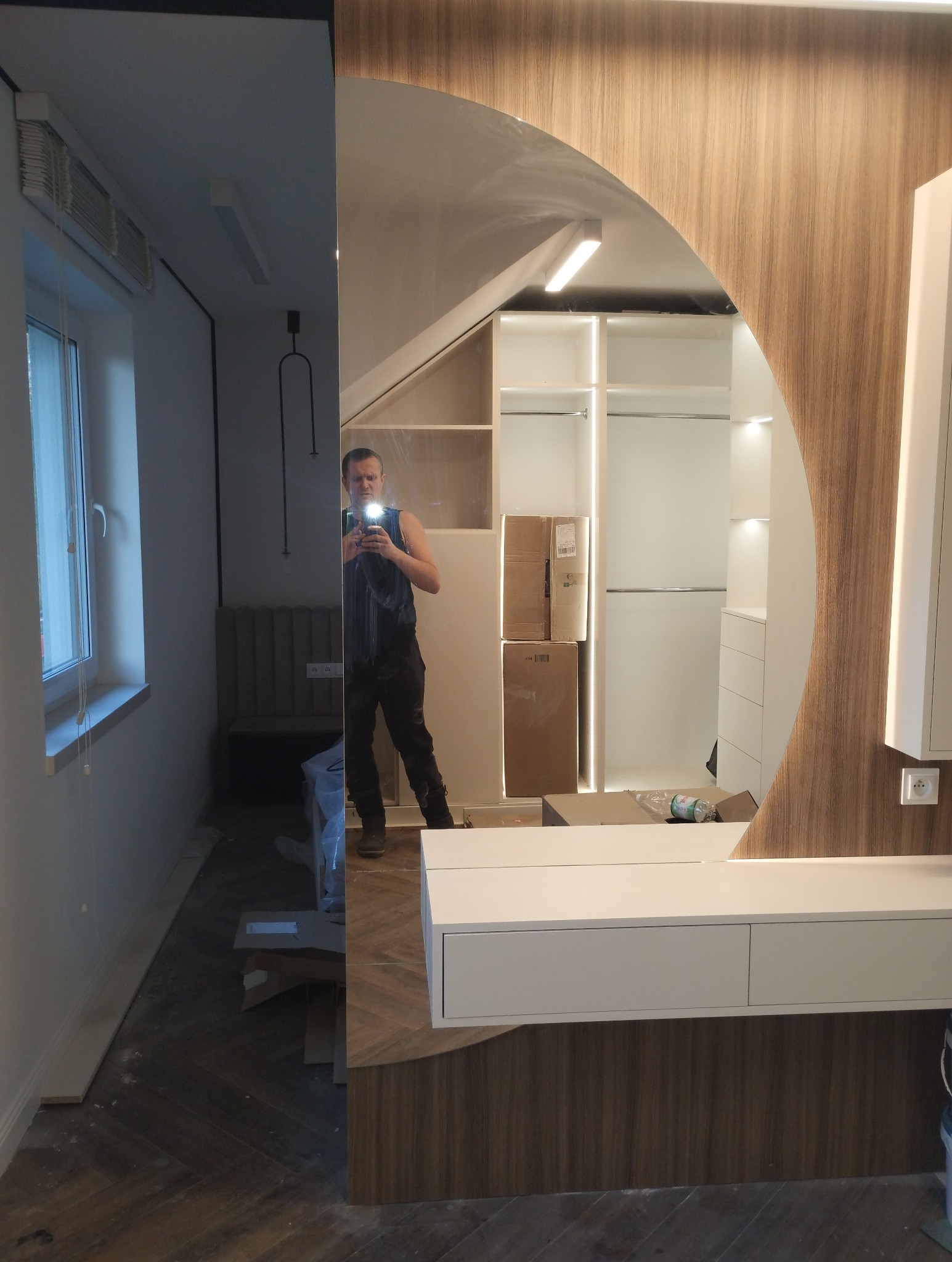 Zamontowane lustro o nietypowym kształcie w zabudowie meblowej, odbijające wnętrze szafy wnękowej z kartonami, widoczne elementy wykończeniowe i odbicie osoby robiącej zdjęcie.