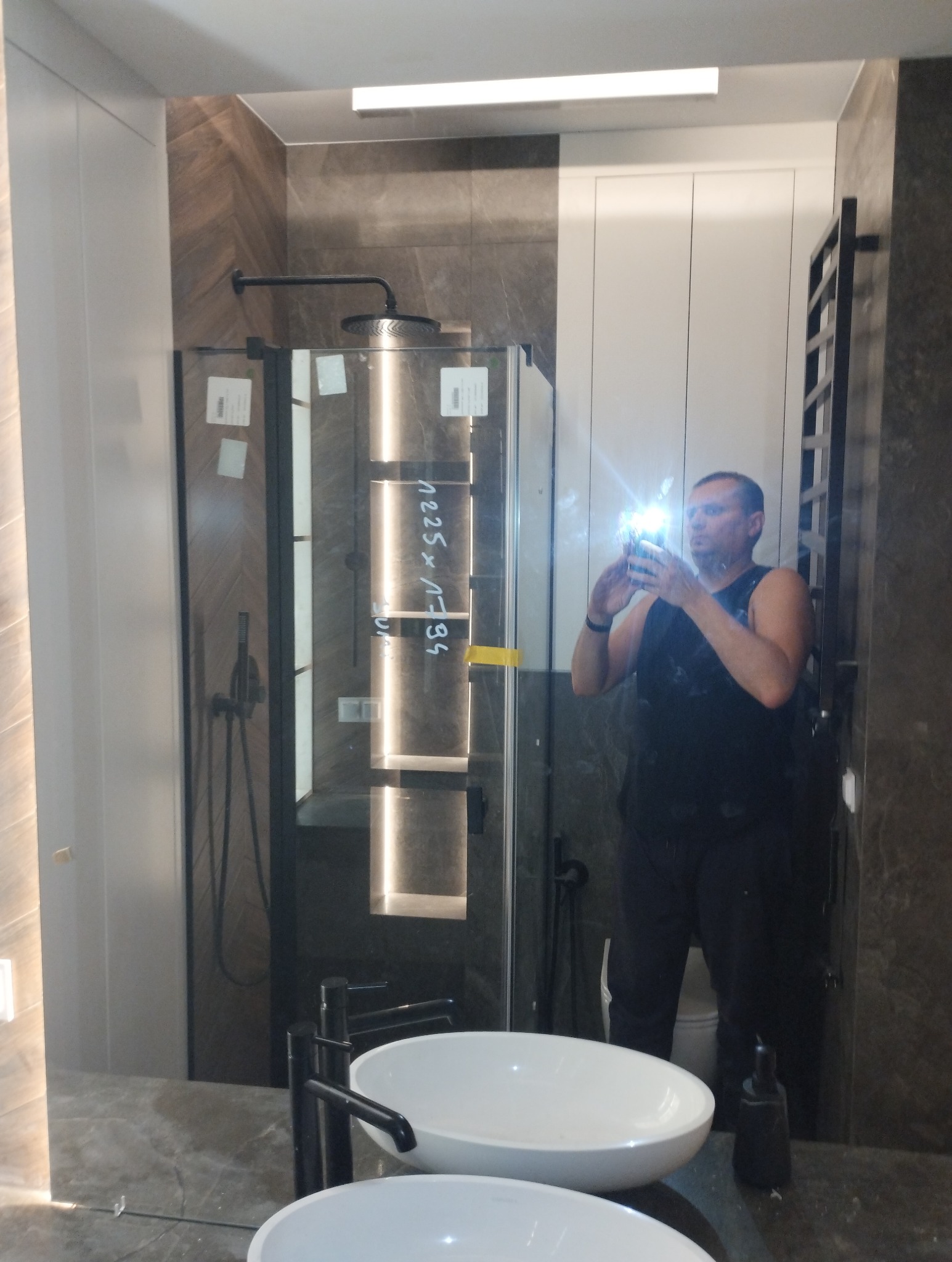 Nowoczesna łazienka z kabiną prysznicową typu walk-in, umywalkami nablatowymi i grzejnikiem drabinkowym, widoczna w odbiciu lustra, z mężczyzną robiącym zdjęcie telefonem.