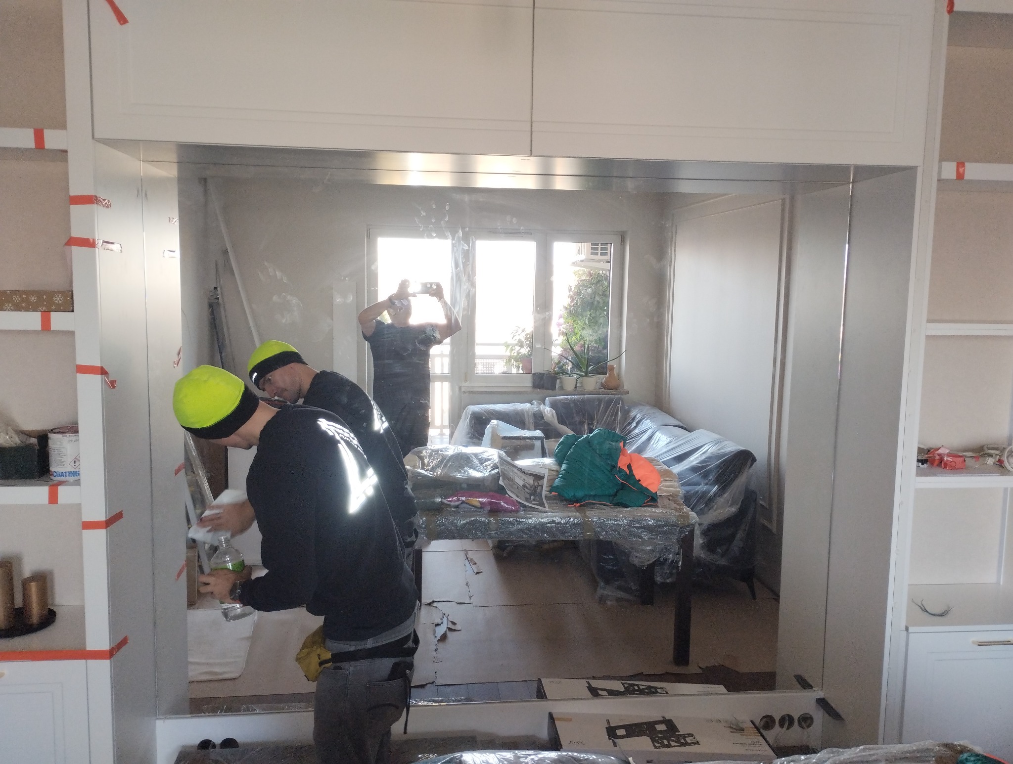 Montaż lustra w zabudowie meblowej przez dwóch pracowników w odblaskowych kamizelkach, jeden czyści powierzchnię, w tle osoba robi zdjęcie telefonem.