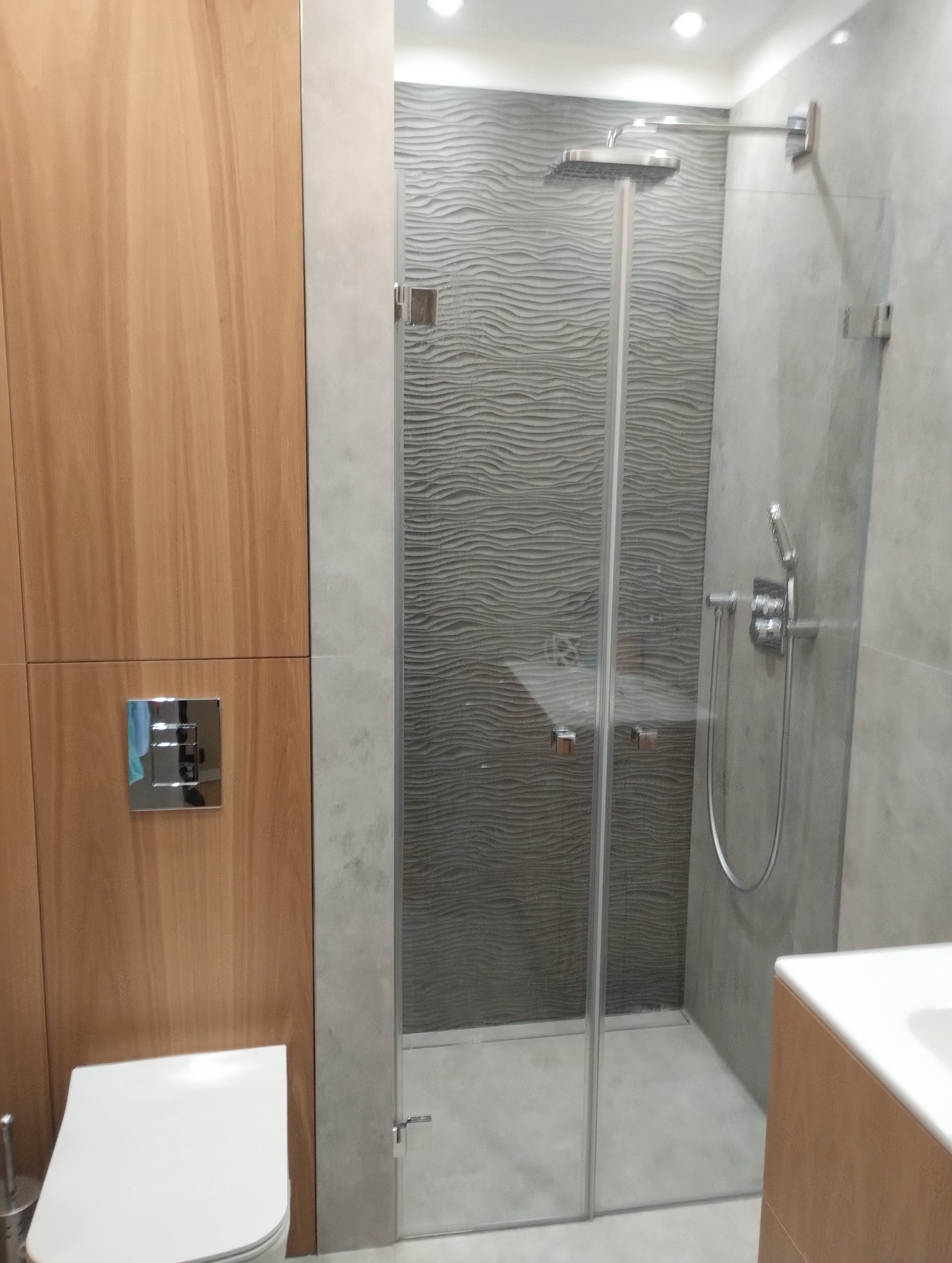 Nowoczesna kabina prysznicowa ze szklanymi drzwiami, deszczownicą i baterią termostatyczną na tle ściany z szarymi płytkami o falistej strukturze, obok drewniana zabudowa toalety.