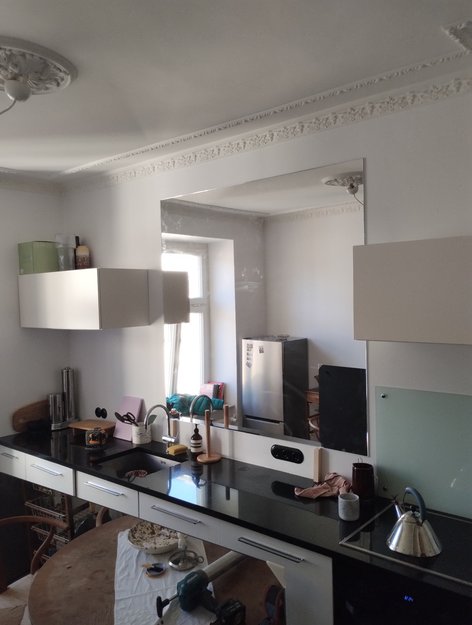 Duże lustro w kuchni, zamontowane na ścianie między szafkami, odbija fragment okna i lodówki; na pierwszym planie blat z czarnym kamieniem i białe szafki, na stole leżą narzędzia.