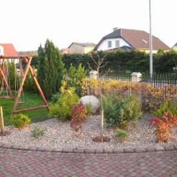 Aranżacja ogrodu z huśtawką, kamieniami ozdobnymi i różnorodną roślinnością. Widoczne krzewy, drzewka i trawnik. W tle domy.