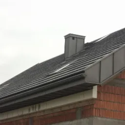 Szary dach z kominem i oknami dachowymi na budynku z czerwonej cegły w pochmurny dzień. Widoczne rynny i podbitka. Detal pokrycia dachowego.