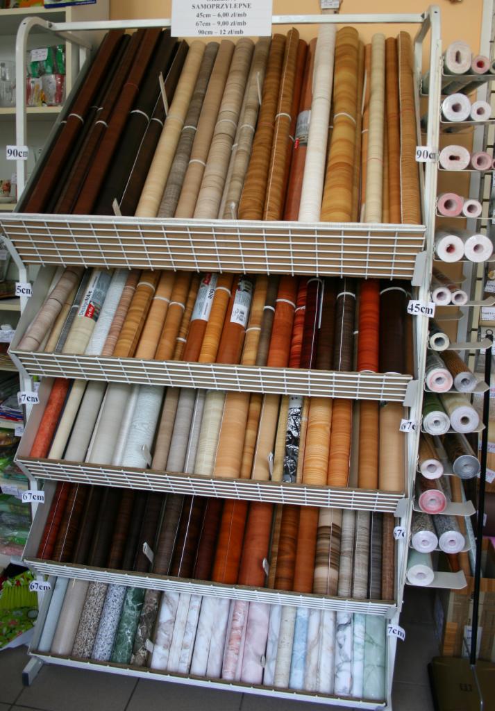Regał w sklepie z okleinami samoprzylepnymi o różnych wzorach i odcieniach drewna, ułożonymi w rolkach na półkach oznaczonych wymiarami.