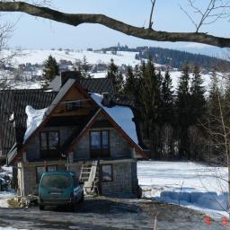 Jan Sikoń F.H.U. SIKONEX - Dom z dachem pokrytym śniegiem w zimowym krajobrazie górskim, z drabina opartą o ścianę, widziany przez gałęzie drzewa.