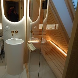 Jan Sikoń F.H.U. SIKONEX - Nowoczesna łazienka z kabiną prysznicową typu sauna, drewniane wykończenie ścian i sufitu w kształcie litery A, biała umywalka na postumencie i złota armatura, oświetlenie LED podkreślające...