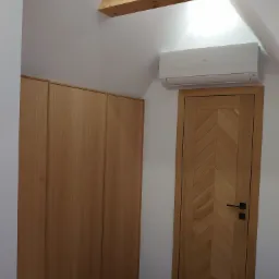 Drewniana zabudowa pod skosem: szafa z gładkimi frontami, drzwi z wzorem jodełki, ozdobna belka z rzeźbieniami i klimatyzator na białej ścianie.