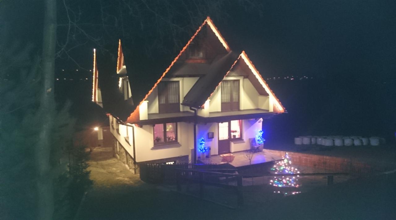 Oświetlony dom z dachem dwuspadowym nocą, dekoracje świąteczne, widok z góry. Czerwone i białe elementy wykończenia. W tle widoczne światła miasta.