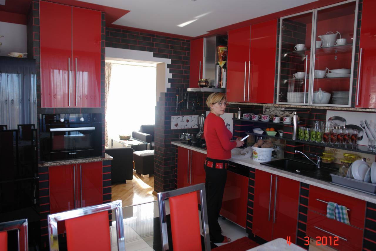 Czerwono-czarna kuchnia z błyszczącymi frontami, wykończona cegłą. Kobieta w czerwonej bluzce stoi przy blacie. Widok na salon przez otwarte przejście.