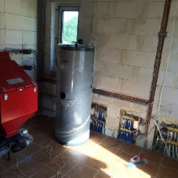 Kotłownia w trakcie montażu: czerwony piec CO, zasobnik na wodę owinięty folią oraz rozdzielacze instalacji CO z rurami w ścianie z betonu komórkowego.