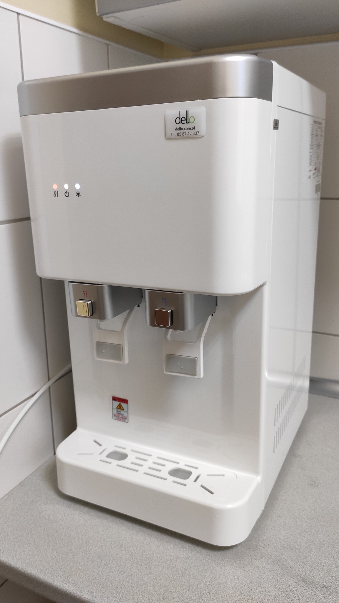 Dystrybutor wody Dello z dwoma kranami, wyświetlaczem LED i tacką ociekową, umieszczony na blacie w kuchni.