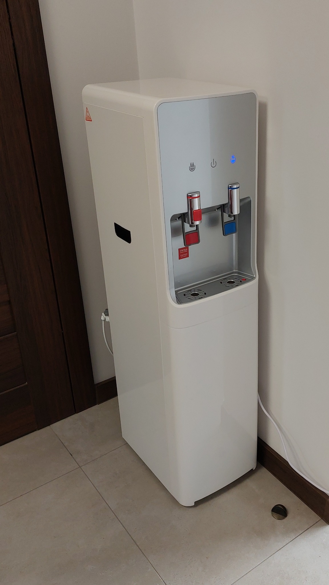 Nowoczesny, biały dystrybutor wody wolnostojący z czerwonym i niebieskim kranem, umieszczony w rogu pomieszczenia z jasnymi płytkami podłogowymi.