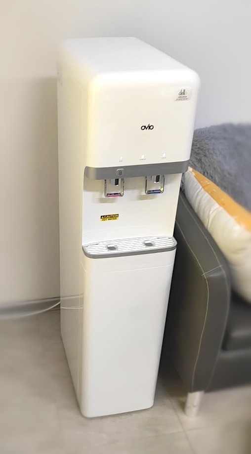 Wolnostojący, biały dystrybutor wody marki ovio z dwoma kranami: czerwonym i niebieskim, umieszczony w biurze obok szarego fotela.