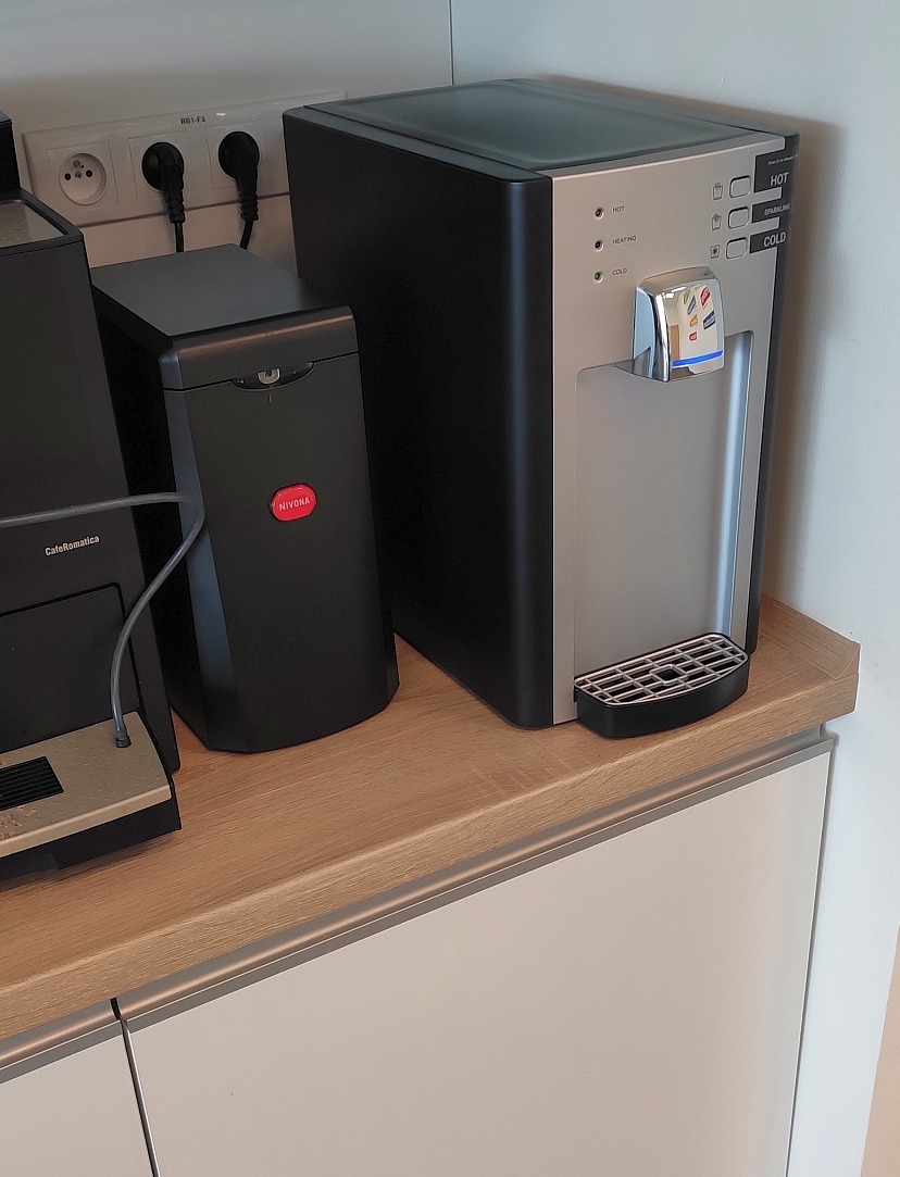 Dystrybutor wody z funkcją wody gazowanej, obok ekspres do kawy, na blacie z jasnego drewna, widoczne gniazdka elektryczne w tle.