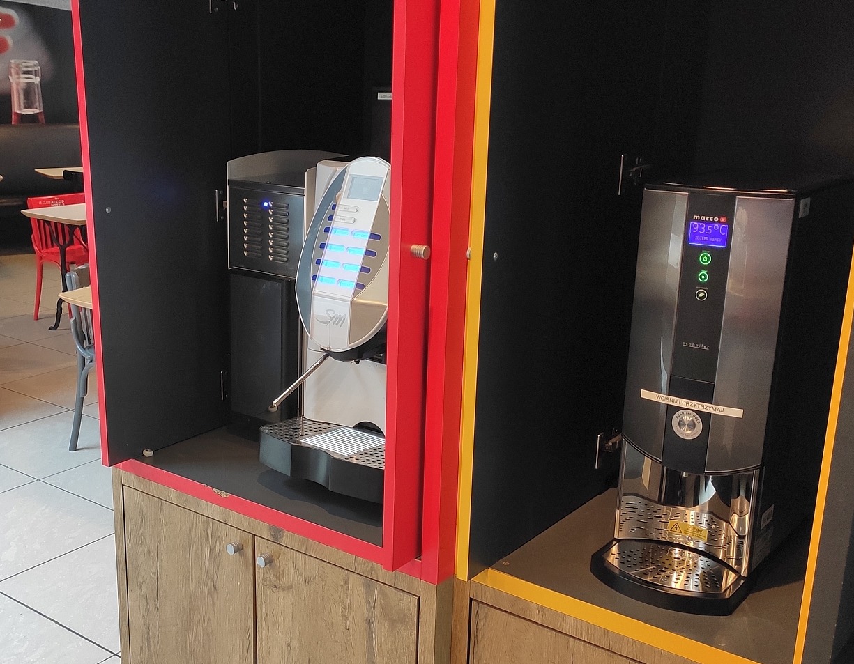 Dwa automatyczne ekspresy do wody w zabudowie meblowej z czerwonymi i żółtymi akcentami, widoczne w tle krzesła w kawiarni.