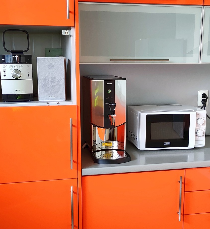Dystrybutor wody Marco obok mikrofalówki na blacie pomarańczowej szafki kuchennej, z widocznym radiem Sony i głośnikiem w otwartej szafce po lewej stronie.